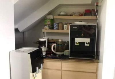Compact communal pantry under a staircase with a coffee machine, water dispenser, and wooden cabinets.