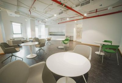 Modern communal breakout area with white cafe tables, green chairs, and exposed ceiling pipes.