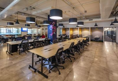 Bright open-plan workspace with long wooden desks and black ergonomic chairs.