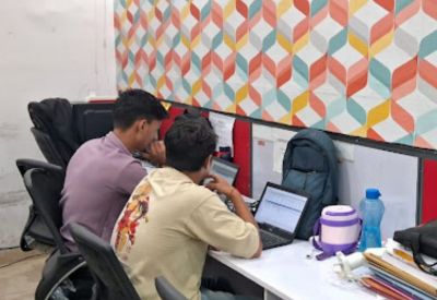 Two people working on laptops at a shared white desk in an office with patterned wallpaper.