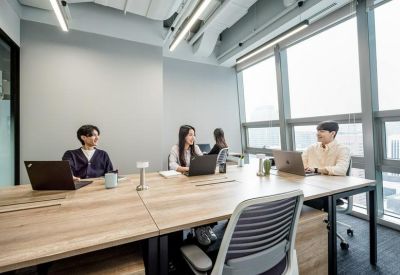 Bright office space with a large wooden communal table and people working on laptops near windows.