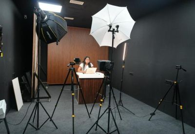 Dark-walled media studio featuring a person at a desk surrounded by cameras and softbox lights.
