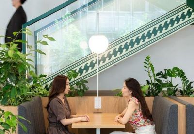 Two women talking at a booth table by a staircase decorated with green patterned glass and plants.