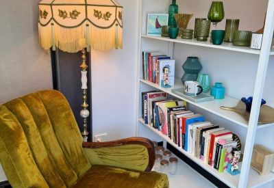 Retro communal lounge featuring a green velvet armchair and white bookshelf with colorful decor.