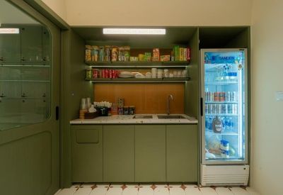 Communal kitchenette featuring green cabinetry, snack shelves, and a transparent beverage fridge.