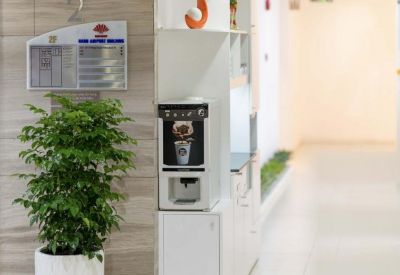 A sleek white self-service coffee station located in a bright hallway next to a large potted plant.