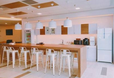 Modern communal kitchen and breakout area with a long wooden island, white stools, and sleek pendant lighting.