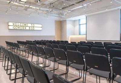 Large presentation room with rows of black chairs facing a projector screen and lightbox sign.