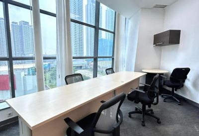 Bright shared workspace featuring a long light-wood desk, ergonomic chairs, and large city-view windows.