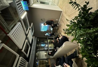 A group of people sitting on a long white bench in a modern lounge area with indoor plants.