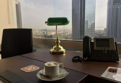 Wooden desk with a classic green banker's lamp and coffee cup overlooking city skyscrapers.