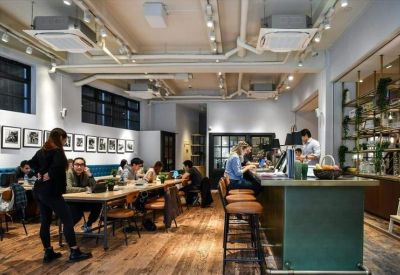 Spacious communal lounge with industrial ceilings, long wooden tables, and people working.