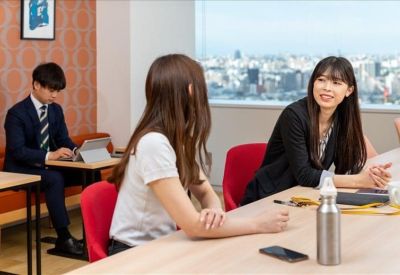 Professionals working and talking in a bright communal lounge with city views and red chairs.
