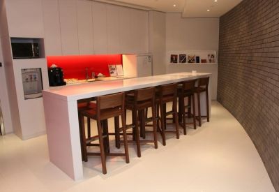 Communal kitchen area with a long white breakfast bar, wooden stools, and vibrant red backsplash.