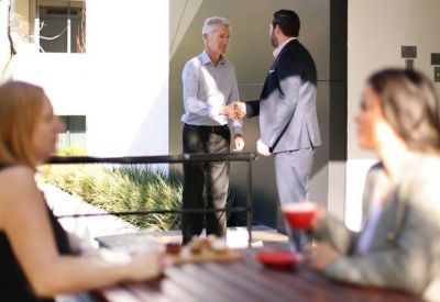 Two businessmen shaking hands in a bright outdoor courtyard area.