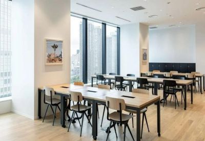 Open-plan workspace featuring long wooden tables and black chairs near large windows.