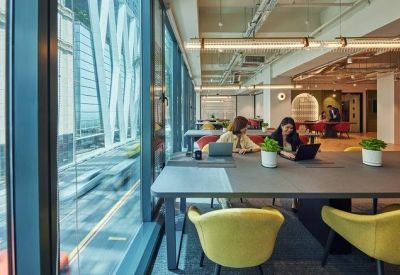 Bright coworking area with long grey tables and yellow armchairs next to large floor-to-ceiling windows.