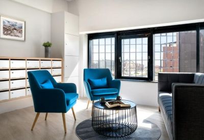 Lounge area with blue velvet armchairs and a circular metal coffee table by large windows.