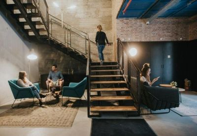 Spacious communal lounge with industrial stairs, teal armchairs, and warm overhead lighting.