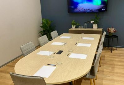 Professional boardroom with a long oval table, grey chairs, and a large wall-mounted screen.