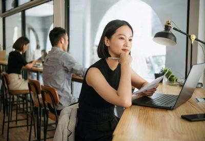 A person working at a wooden desk with a laptop and lamp in a bright coworking space.