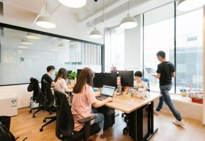 Bright open-plan office with people working at wooden desks near large windows.