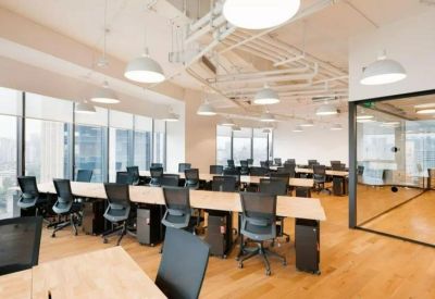 Bright open-plan office with long wooden desks, ergonomic chairs, and large windows.