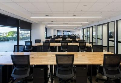 Open-plan office with rows of wooden desks, black ergonomic chairs, and river views through windows.