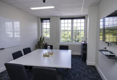 Sunlit meeting room with a large white square table, black chairs, and windows overlooking greenery.
