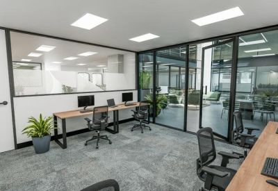 Modern office space with workstations, glass partitions, and a grey carpeted floor.
