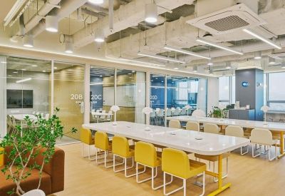 Open-plan workspace with long white communal tables, yellow chairs, and glass-walled meeting rooms.