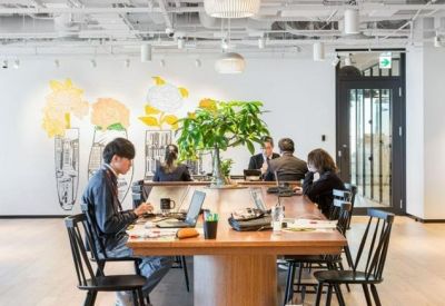 Bright coworking area with people working at a long wooden table decorated with large green plants.