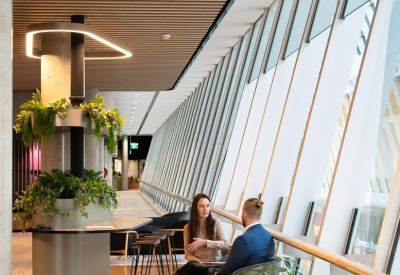 Sunlit communal lounge area with people sitting by floor-to-ceiling windows and indoor greenery.