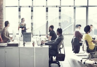 Professional open-plan office space with employees working at white desks against a brick wall.