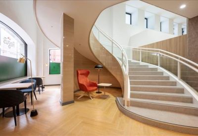 Elegant curved staircase in a light-filled lobby area with wooden flooring and a red accent chair.