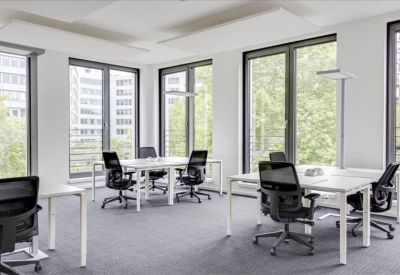 Sunlit open-plan office with white desks, black chairs, and large windows overlooking trees.