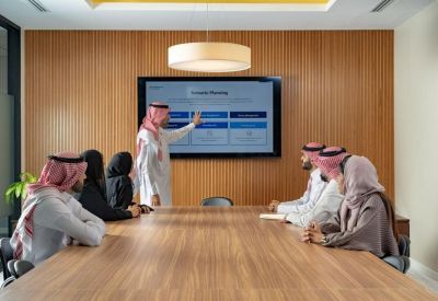 Boardroom meeting in progress with people seated around a long wooden table in front of a screen.