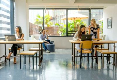 Bright open-plan coworking space with people working at wooden tables by large windows.