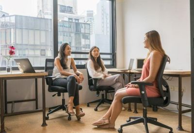 Three women having a casual meeting in an office lounge area with comfortable black chairs.