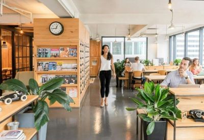Spacious sunlit lounge with a large wooden bookshelf wall and people working in the background.