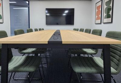 Large boardroom with a long light-wood table, green upholstered chairs, and a wall-mounted screen.