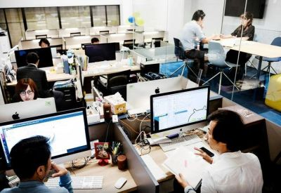 Busy open-plan office with professionals working at computer workstations in a bright environment.