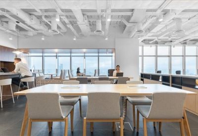 Open-plan office space with long white desks and views of the city skyline through large windows.