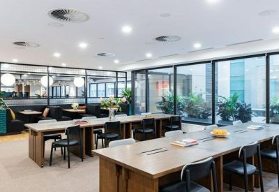 Bright coworking floor featuring long communal wooden desks and black ergonomic chairs.