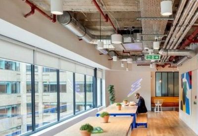 Bright open-plan workspace with long wooden tables and exposed ceiling pipes.