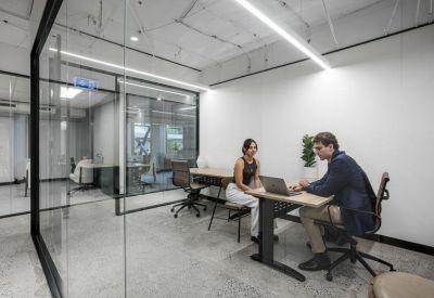 Modern glass-partitioned office with two people working at a wooden desk.