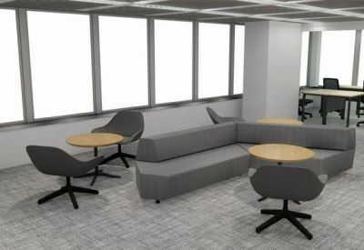 Lounge area with a grey sectional sofa, wooden side tables, and views of the office workspace.