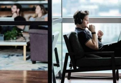 A man wearing headphones works on a laptop in a modern breakout space with large windows.