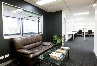 Lounge area with a brown leather sofa, glass coffee table, and an open-plan office in the background.
