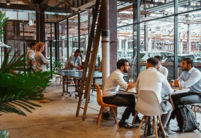 Sun-drenched breakout space with groups of people working at tables near large glass windows.